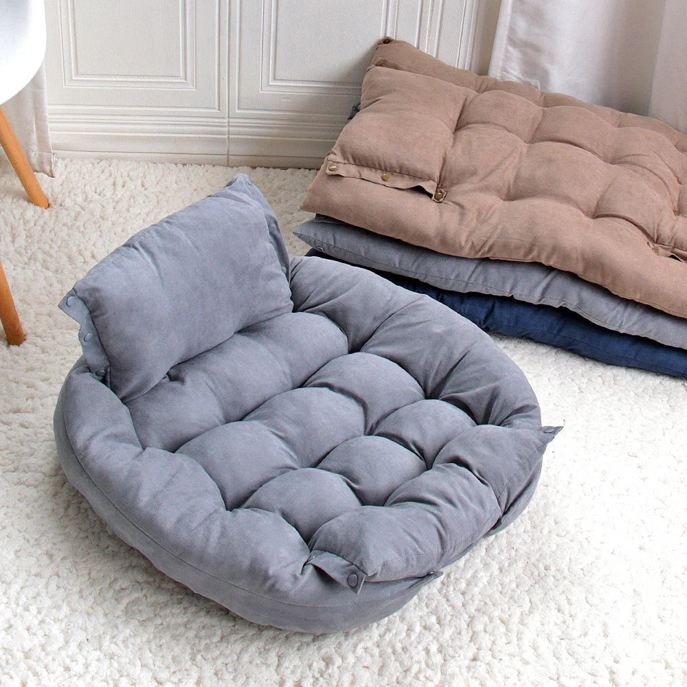 Grey pet bed on a white carpet with folded blankets in the background